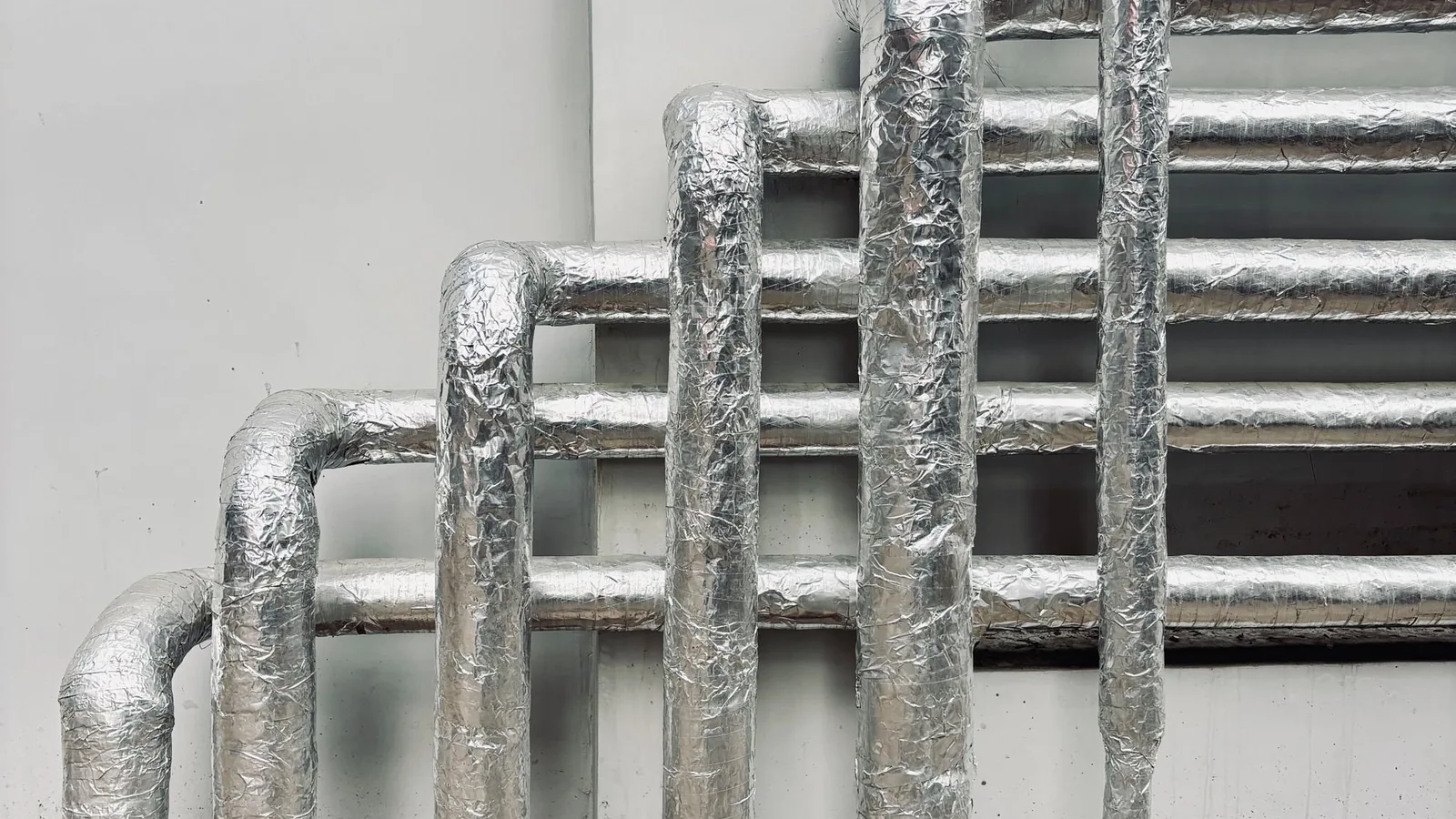 Foil-insulated pipes run mini split line set through exterior wall, wrinkled metallic silver insulation on white surface